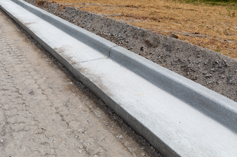 Concrete curb installation alongside a road under construction, showing a smooth finished curb and adjacent soil