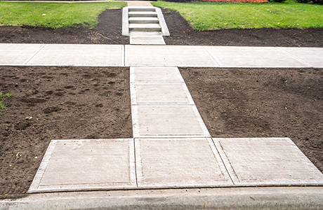 Newly installed concrete sidewalk with stairs leading to a residential area, surrounded by soil and green landscaping