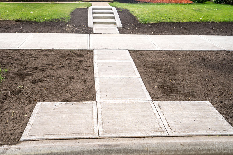 Newly installed concrete sidewalk with stairs leading to a residential area, surrounded by soil and green landscaping