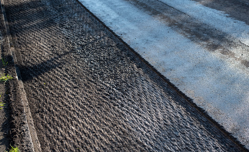 Close-up of asphalt milling process removing the surface layer of the road, showing textured grooves and prepared surface for repaving